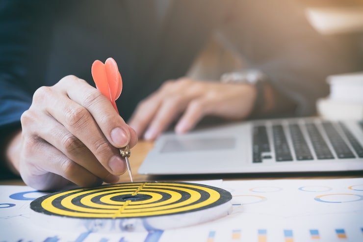 A close-up of a person in a suit holding a red dart precisely over the center of a small yellow and black bullseye target on a desk.